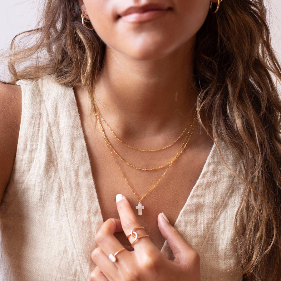 a mother of pearl white cross pendant on a 14k gold fill chain, photographed on a model wearing a cream linen vest and other 14k gold fill chain necklaces made by Token Jewelry