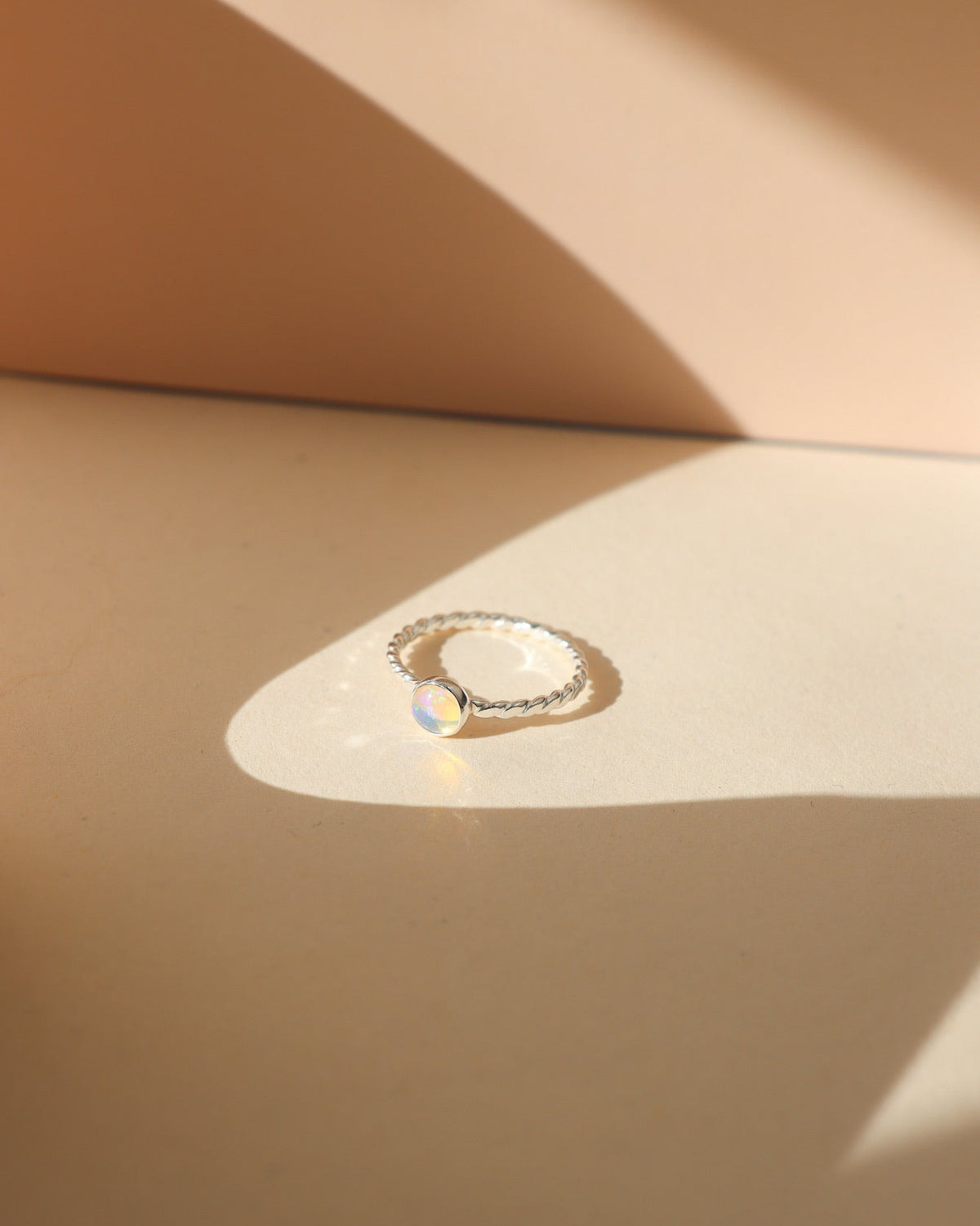 Silver twisted-band ring featuring a luminous round opal gemstone with iridescent flashes of pink, green, and yellow, photographed on a warm cream surface with soft natural sunlight and sculptural shadows.