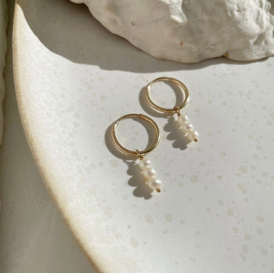 14k gold fill Petite Pearl Hoops laid on a white plate in the sunlight. These earrings feature a Goldie hoop with three tiny gemstone studs. - Token Jewelry