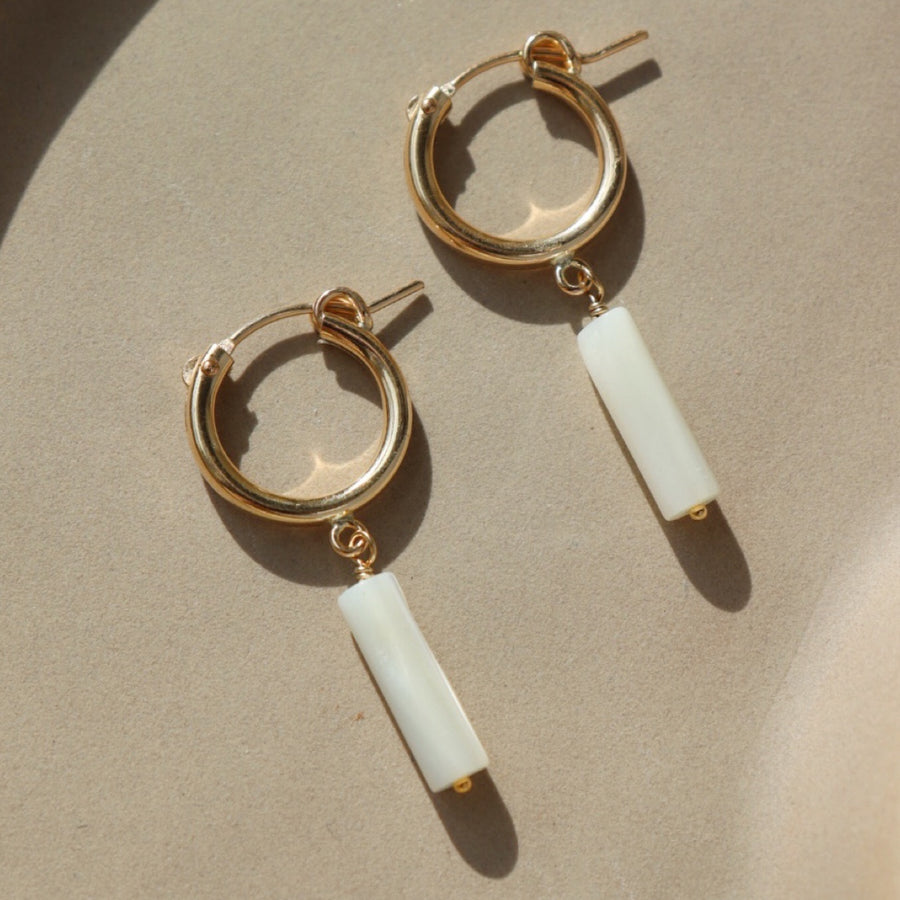 14k gold fill Mother of Pearl Hoops laid on a peach plate in the sunlight. These earring feature a classic hoop with a mother of pearl drop down stone.