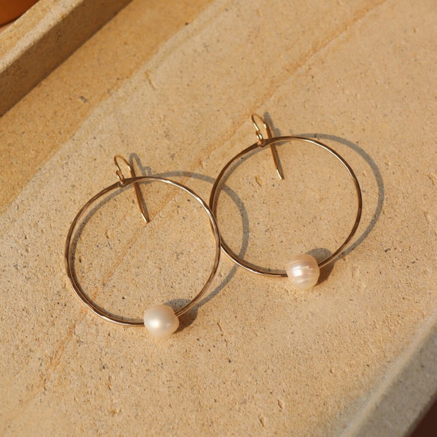 14k gold fill Pearl Hoops laid on a white plate in the sunlight. These earrings feature a hammered hoop with a pearl gemstone connecting the hoop together.