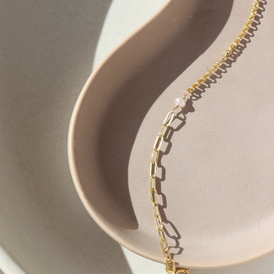 14k gold fill bracelet place on top of a peach colored plate. Sitting in the sunlight, featuring the narrow link chain, a tiny pearl gemstone, and the Dylan chain. The jewelry is handmade in Eau Claire Wisconsin, at Token Jewelry.