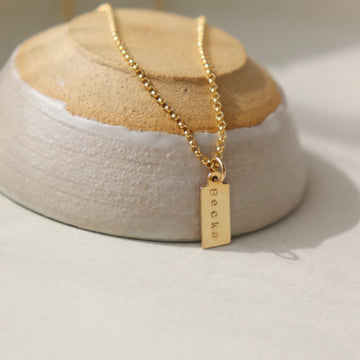 14k gold fill Mini Tag Personalized Necklace placed on a tan and gray bow. This necklace features the coin necklace chain followed by a mini tag with personalization of your choice.