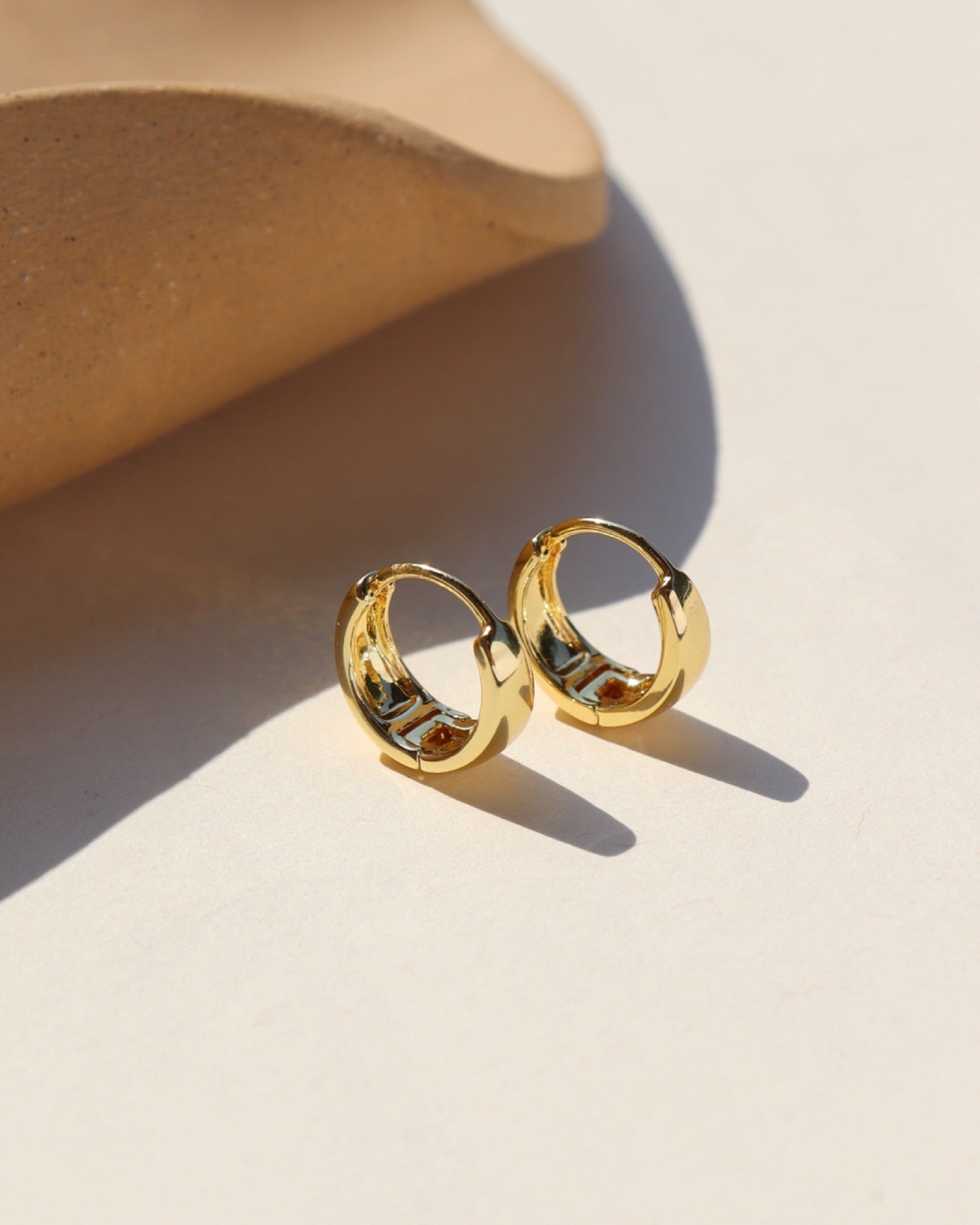 Small, polished gold filled hinged hoop earrings sitting near a ceramic dish in the sunlight. 